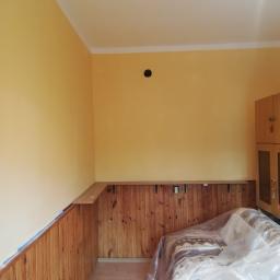 usługi malarskie Marcel - Pokój w trakcie malowania: żółte ściany, drewniana boazeria zabezpieczona niebieską taśmą malarską, meble przykryte folią ochronną.