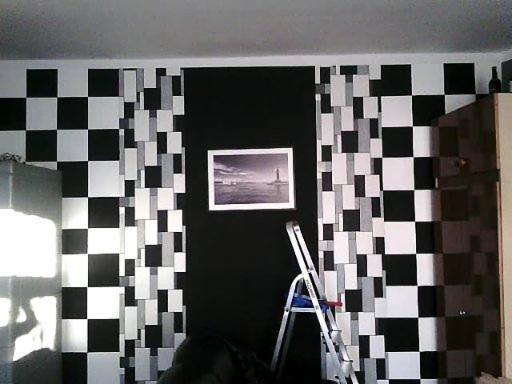 Ściana pomalowana w czarno-białą szachownicę z pionowymi pasami mozaiki z prostokątów, na czarnej ścianie wisi obrazek z latarnią morską, przy ścianie stoi drabina.