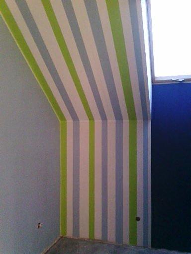 Pomalowane ściany w pokoju dziecięcym w pionowe, zielono-niebieskie pasy. Skośny sufit pomalowany w ten sam wzór. Fragment okna w prawym górnym rogu.