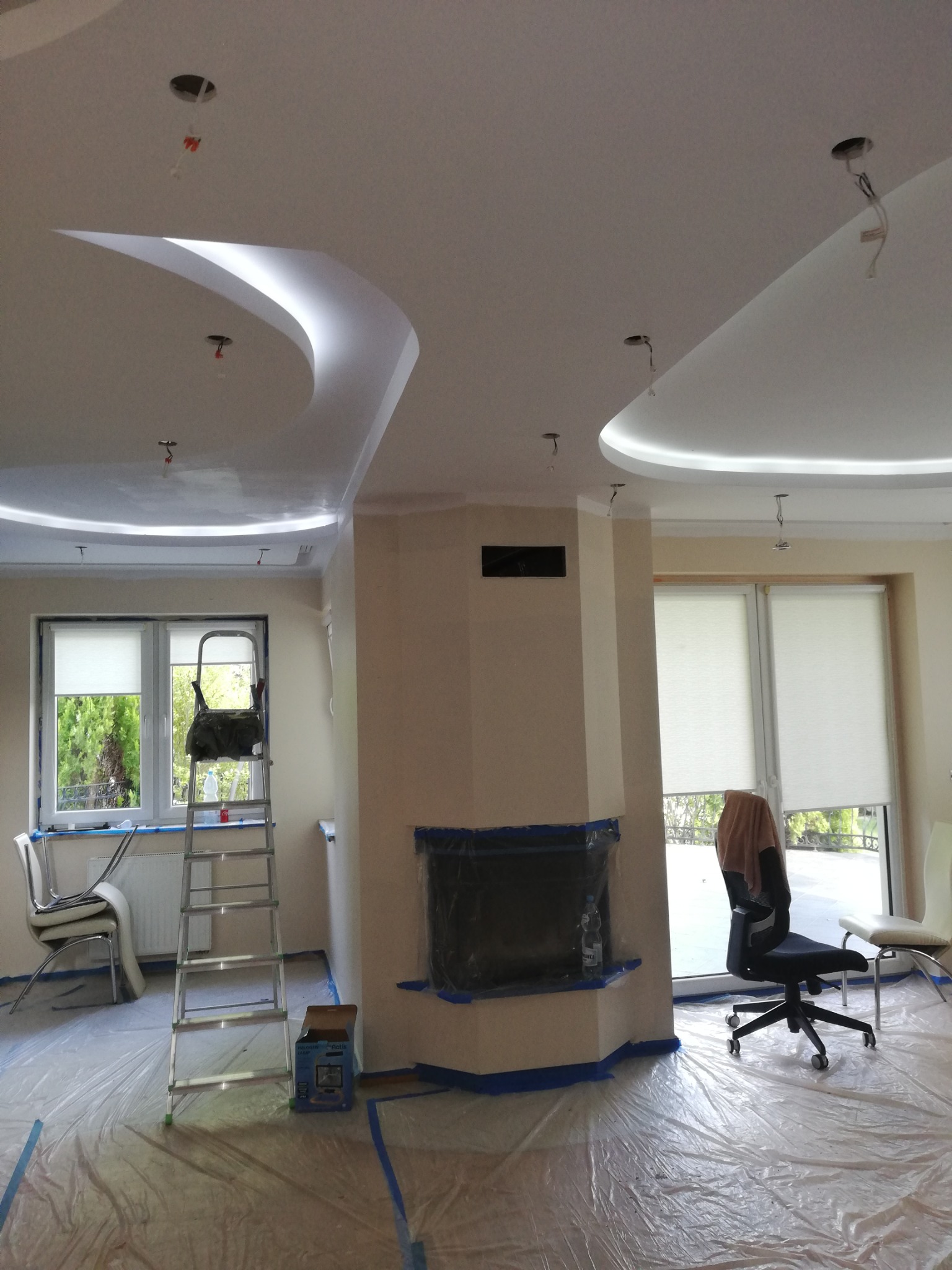 Pokój w trakcie prac wykończeniowych: sufit z dekoracyjnymi elementami oświetleniowymi, zabezpieczona folią podłoga, drabina, zaklejony taśmą kominek, przygotowane do pracy pomieszczenie.