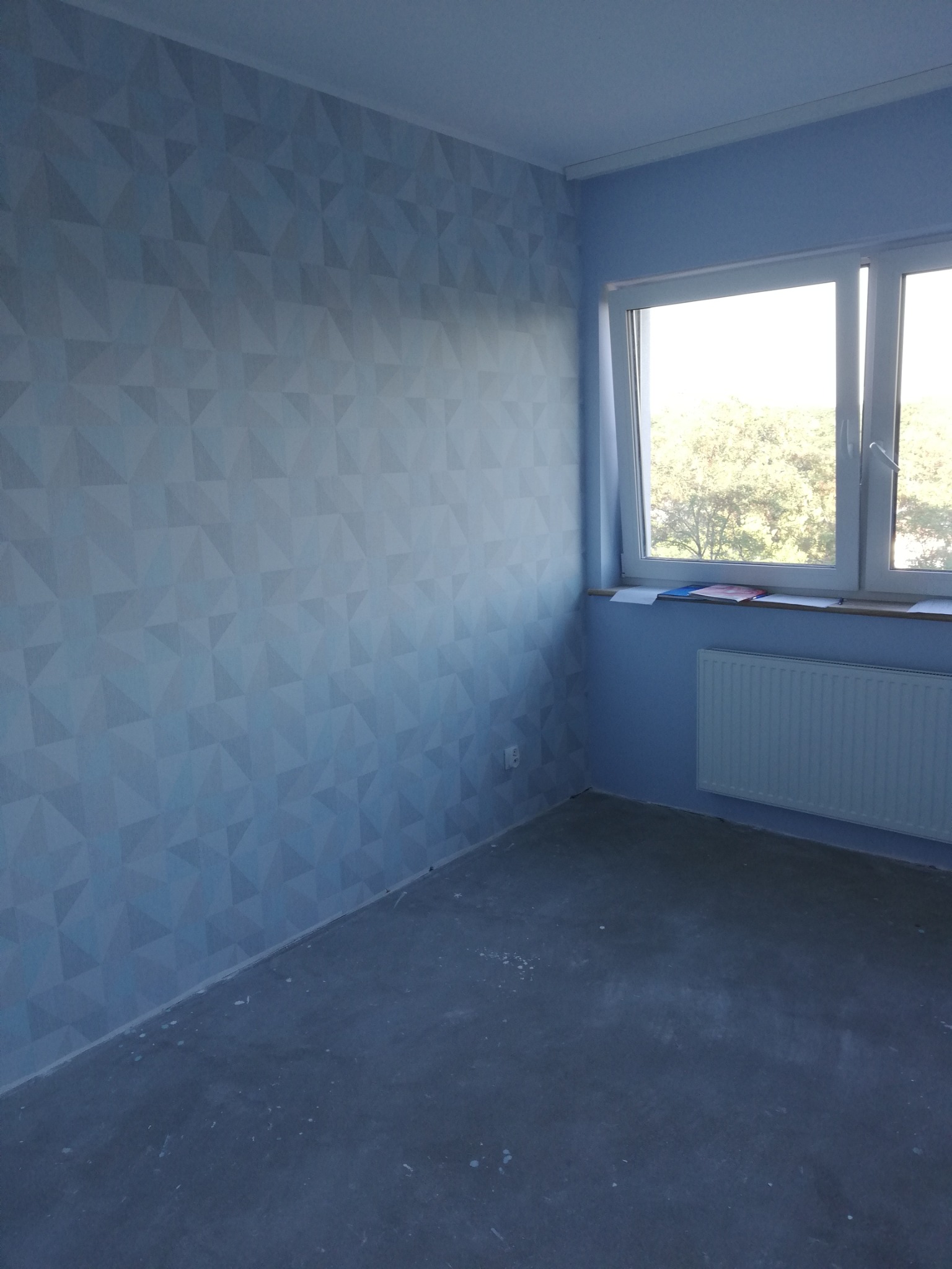 Pomalowane na niebiesko ściany pokoju z geometrycznym wzorem trójkątów, okno z białymi ramami i grzejnik pod nim, surowa podłoga.