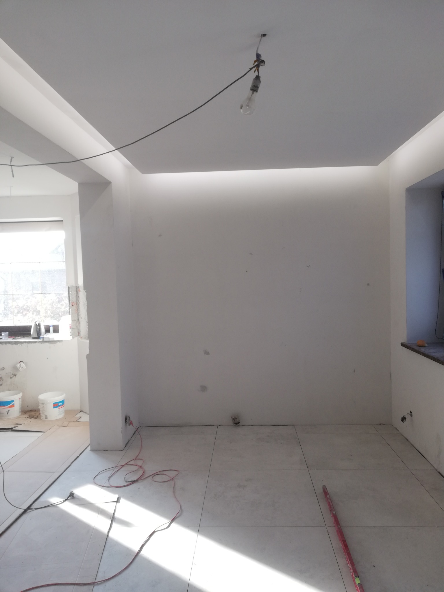 Pokój w trakcie wykańczania, świeżo pomalowane ściany na biało, widoczne ślady po gładzi, na podłodze płytki, kabel zwisający z sufitu z żarówką, w tle wiaderka z farbą.
