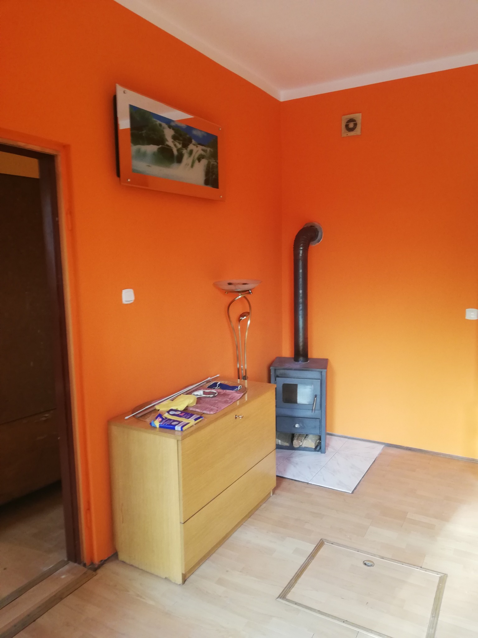 Pomarańczowe ściany w pokoju po remoncie, widoczny piec wolnostojący z rurą, komoda z akcesoriami malarskimi oraz obrazek na ścianie.