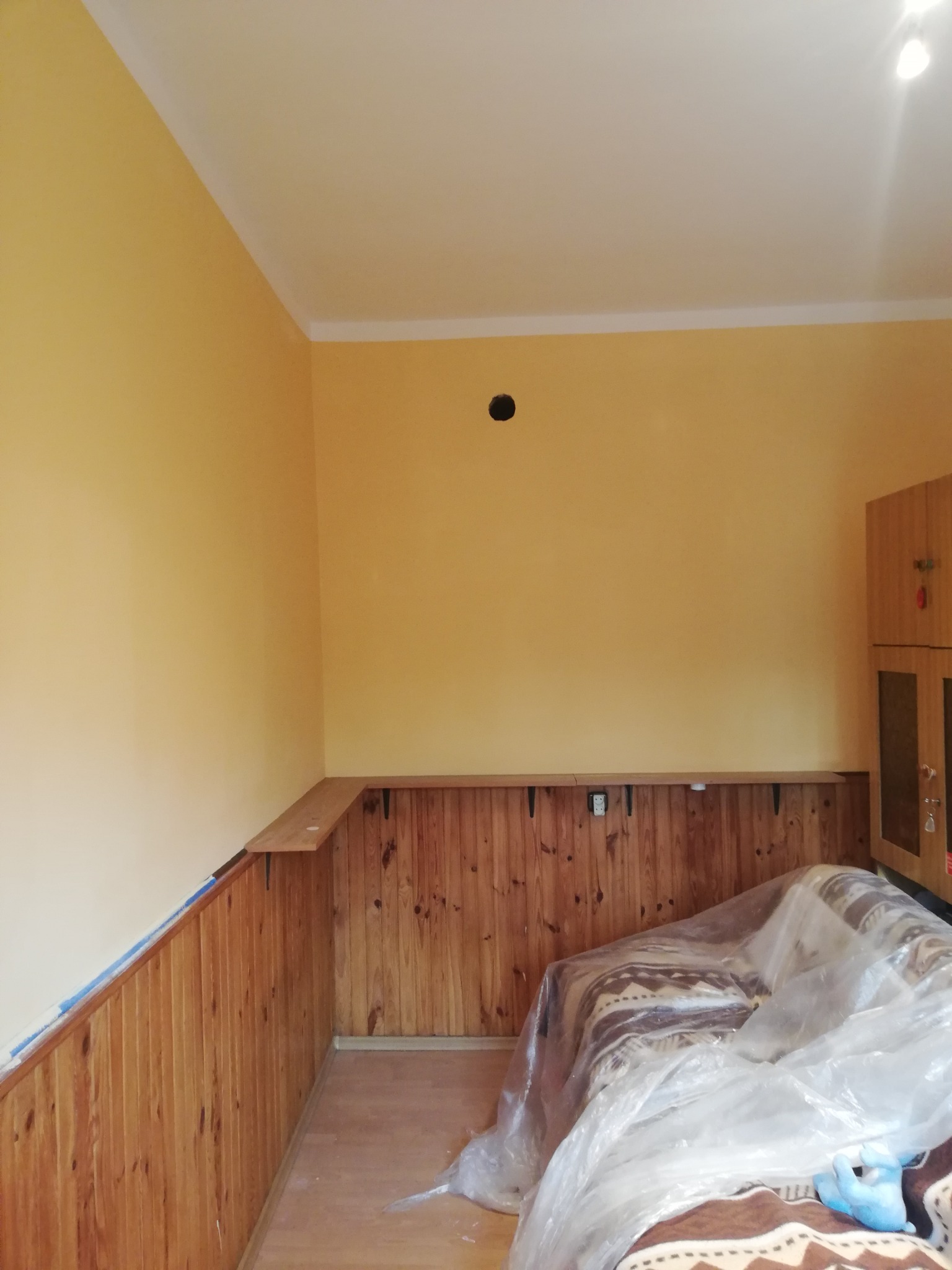 Pokój w trakcie malowania: żółte ściany, drewniana boazeria zabezpieczona niebieską taśmą malarską, meble przykryte folią ochronną.