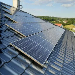 Panele słoneczne na dachu z czarnej dachówki ceramicznej, widok z góry, w tle drzewa i zabudowania.