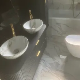 Nowoczesna łazienka z marmurowymi płytkami, umywalkami nablatowymi w odcieniach bieli i złota armatura. Minimalistyczny design i eleganckie wykończenie.