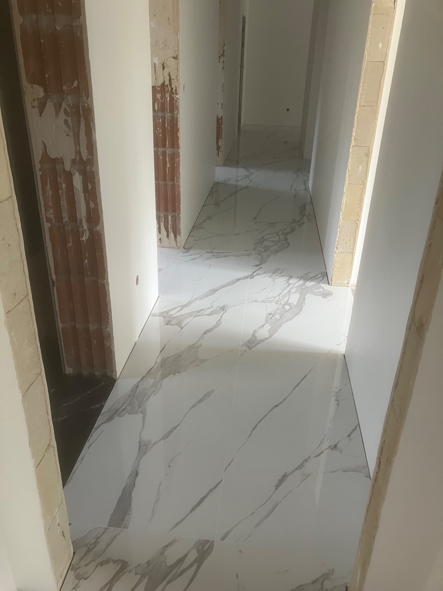 Biała marmurowa podłoga z szarymi żyłami w korytarzu w trakcie wykończenia. Widoczne surowe ściany i przygotowane do malowania, z elementami cegły i betonu.