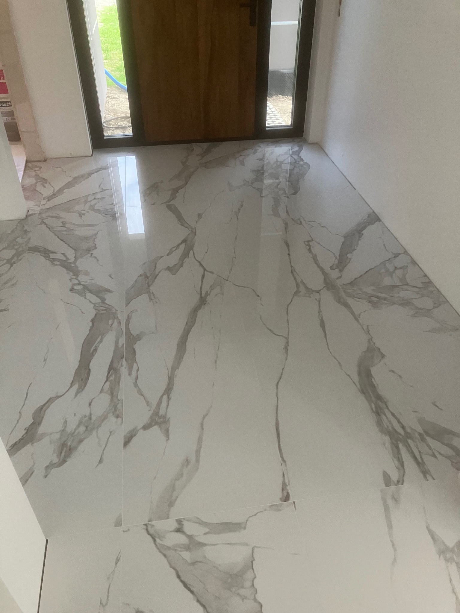 Połyskujące, białe płytki podłogowe z szarym wzorem żyłkowania, ułożone w nowym pomieszczeniu z drewnianymi drzwiami. Widok z góry.