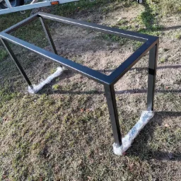Czarna, metalowa konstrukcja stołu z profili kwadratowych, zabezpieczona folią na nóżkach, leżąca na trawie, widoczny fragment przyczepki samochodowej w tle.