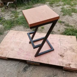 Nowoczesny stołek barowy z drewnianym siedziskiem i metalową ramą w kształcie litery Z, ustawiony na płycie OSB w otoczeniu trawy i betonu.