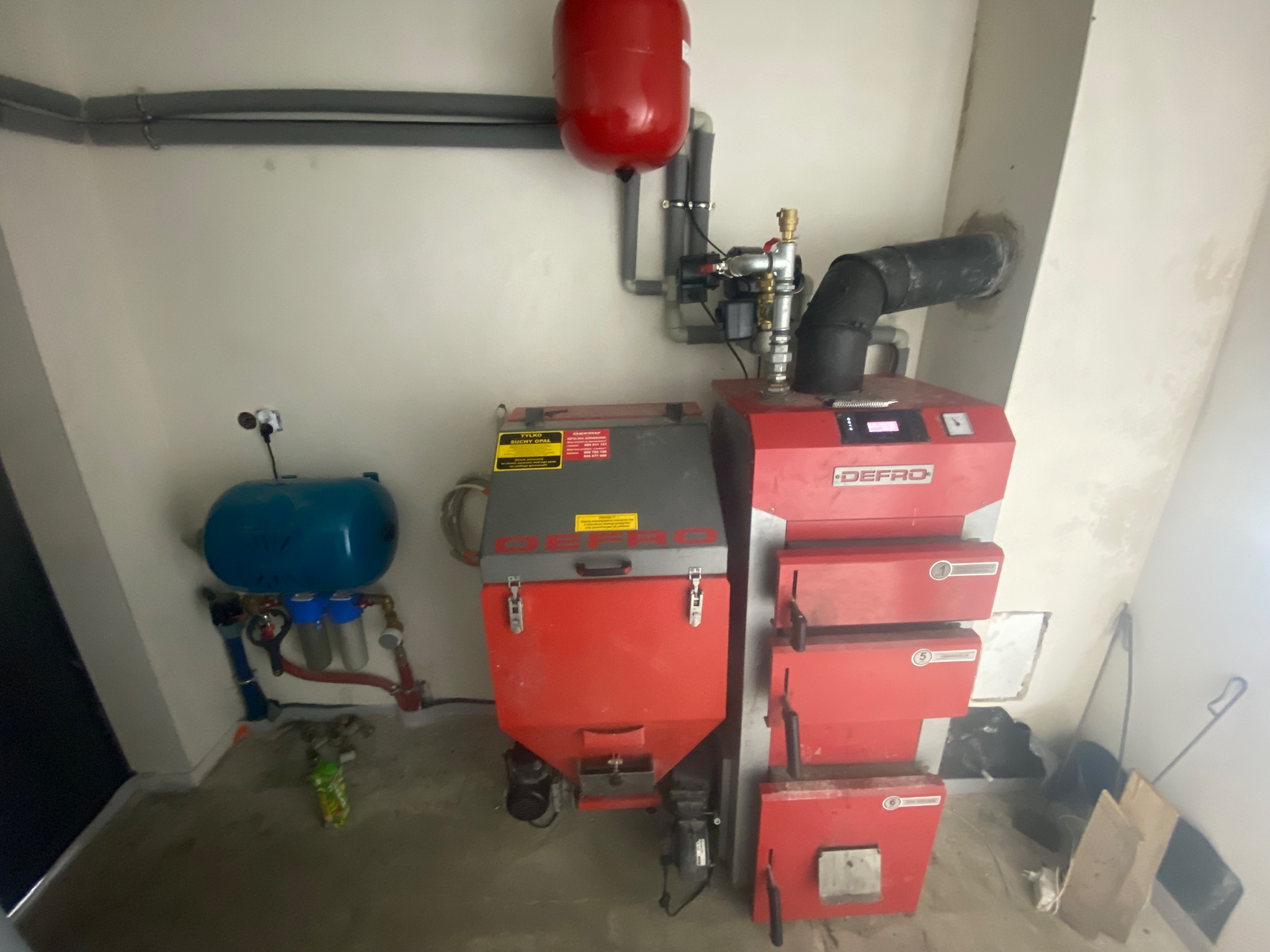 Czerwono-szary piec Defro z podłączonymi instalacjami w kotłowni. Widoczne rury, zbiornik wyrównawczy i filtry. Ściany białe, surowy stan wykończenia.