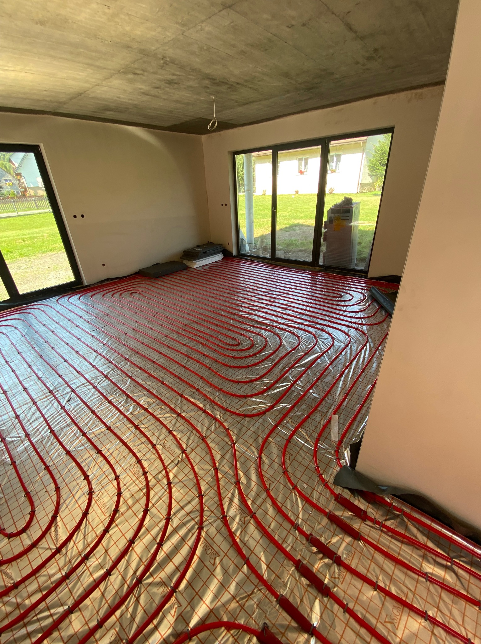 Czerwone rury ogrzewania podłogowego ułożone na folii w surowym wnętrzu z dużymi oknami i betonowym sufitem. Widok na zewnątrz przez okna.