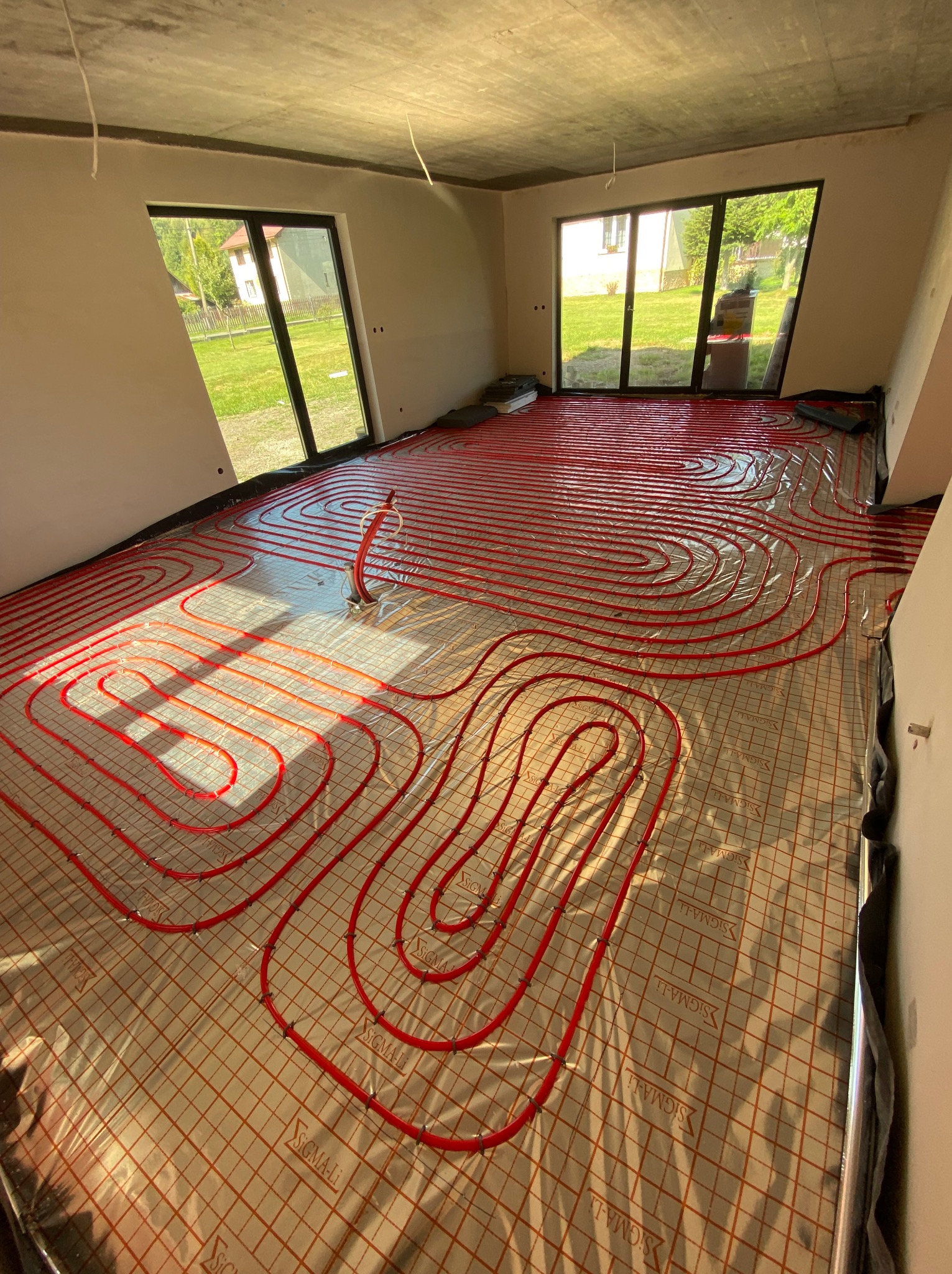 Czerwone rury ogrzewania podłogowego ułożone spiralnie na folii z nadrukiem w nowym, jasnym pomieszczeniu z dużymi oknami i surowym betonowym sufitem.