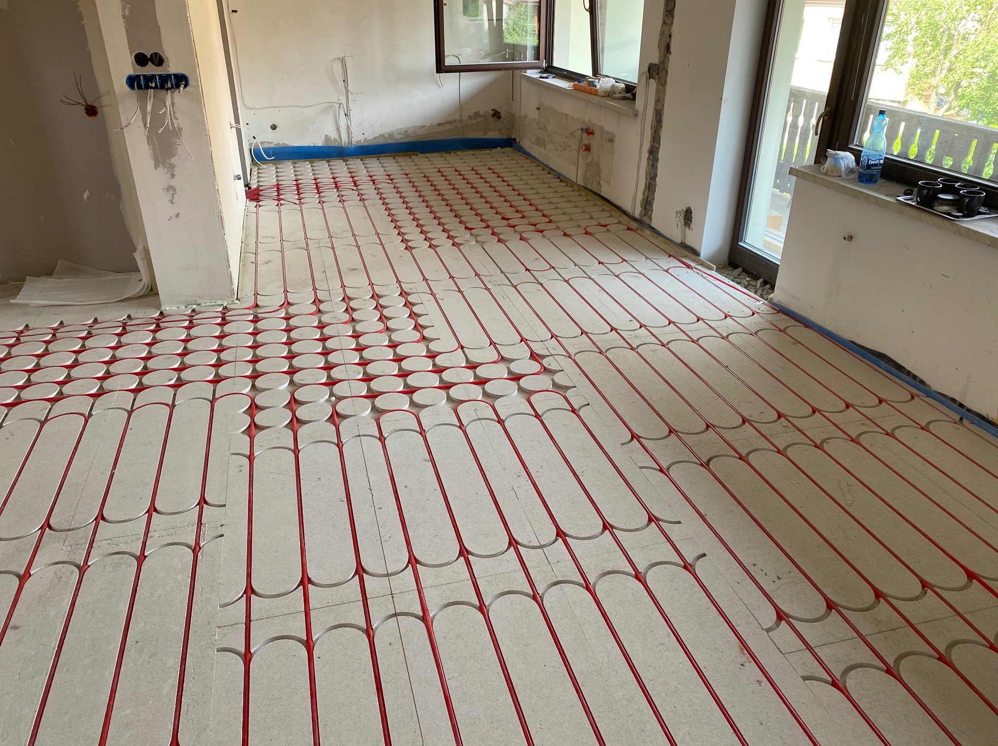 Czerwone rurki ogrzewania podłogowego ułożone na białych płytach izolacyjnych w surowym wnętrzu. Widoczne okno i instalacje elektryczne w ścianie.