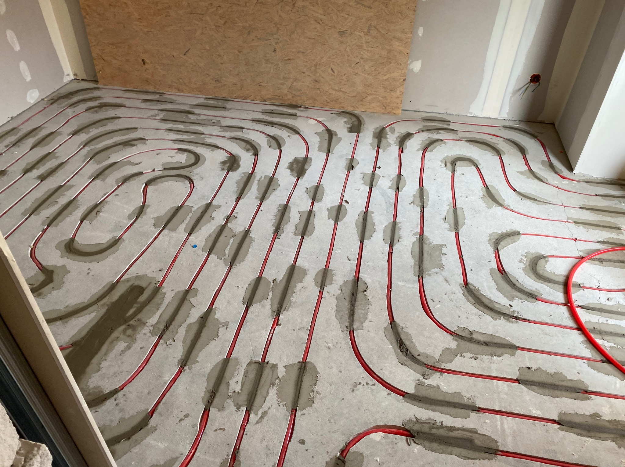 Czerwone rury ogrzewania podłogowego ułożone w spiralny wzór na szarym betonie, częściowo zalane zaprawą. Widok z góry, surowy stan wykończenia wnętrza.
