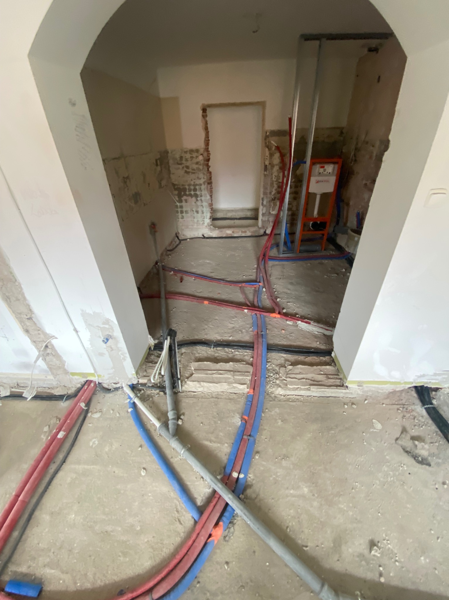 Instalacja hydrauliczna w trakcie remontu: widoczne rury w kolorach czerwonym i niebieskim, prowadzone po podłodze w surowym stanie. W tle stelaż podtynkowy WC.