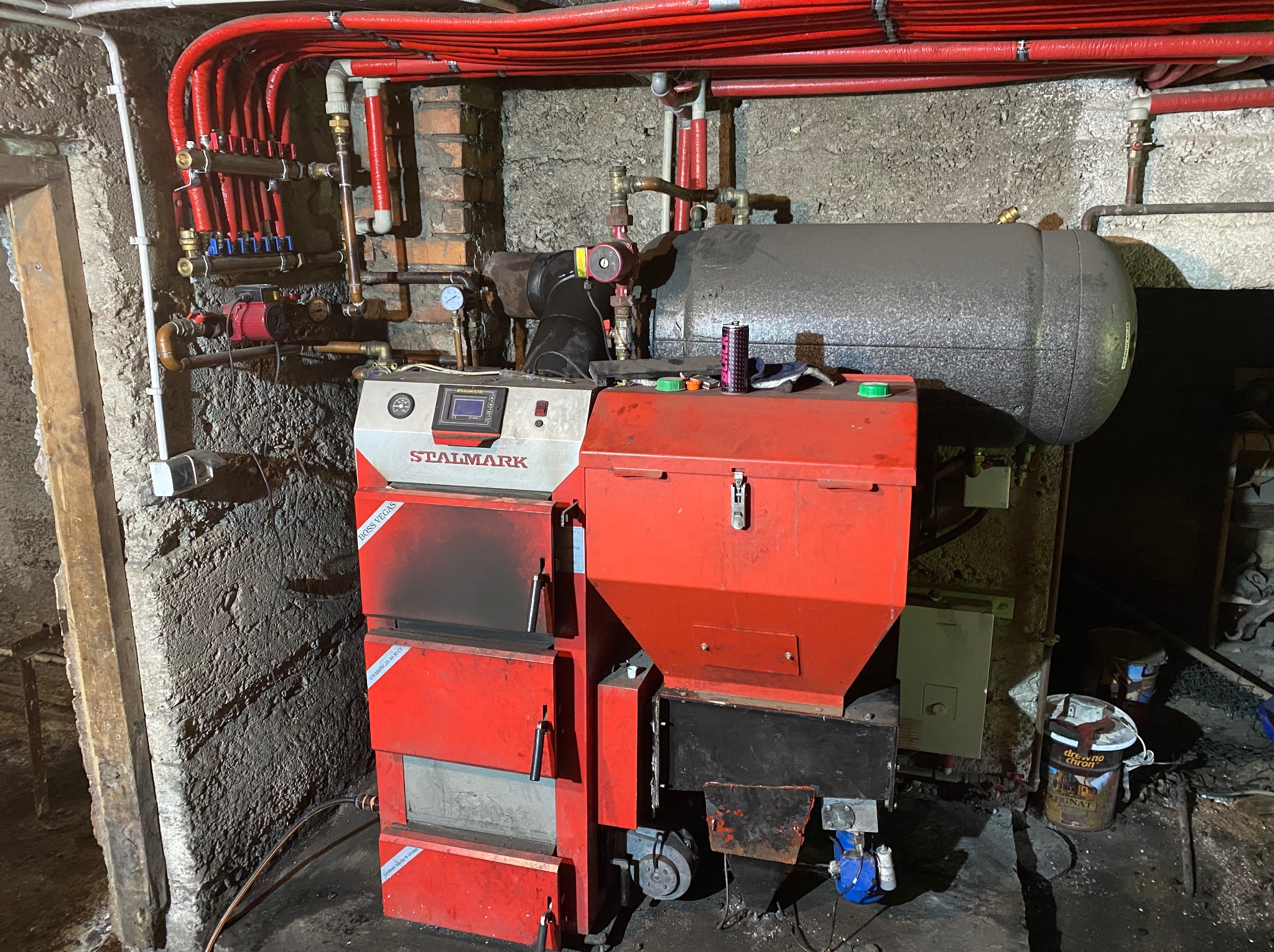 Czerwony piec CO marki Stalmark z podłączoną instalacją grzewczą w kotłowni o surowych ścianach. Widoczne rury i zbiornik wyrównawczy.