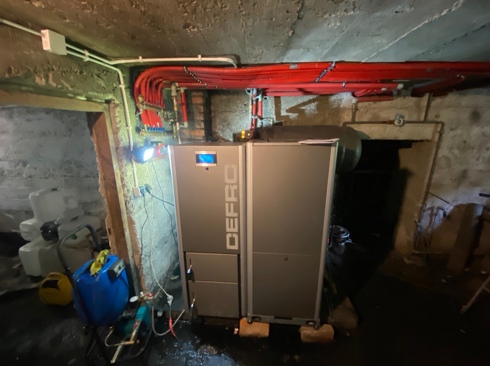Nowoczesny piec DEFRO w piwnicy z instalacją grzewczą z czerwonych rur. Widoczne zbiorniki i narzędzia hydrauliczne. Oświetlenie punktowe.