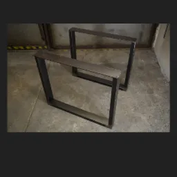 Dwie stalowe, spawane ramy w kształcie prostokąta, prawdopodobnie nogi do stołu, na tle betonowej podłogi i ściany z widocznymi rurami.