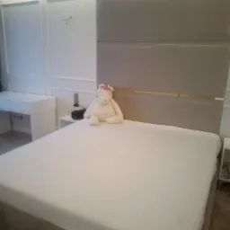 Beżowe łóżko z tapicerowanym zagłówkiem w nowoczesnej sypialni z białymi ścianami i kryształowym żyrandolem, na łóżku siedzi pluszowy jednorożec.