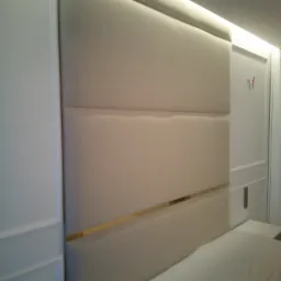 Tapicerowany zagłówek łóżka w odcieniach bieli z poziomymi, złotymi akcentami, wkomponowany w klasyczną, białą boazerię, oświetlony od góry subtelnym światłem LED.