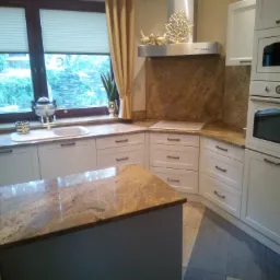 Jasna kuchnia z granitowym blatem, wyposażona w białe szafki z poziomymi uchwytami, piekarnik i mikrofalówkę w zabudowie oraz wyspę kuchenną z blatem o podobnym wzorze.