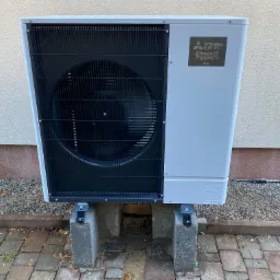 Zewnętrzna jednostka pompy ciepła Mitsubishi Zubadan R32, zamontowana na betonowych wspornikach przy ścianie budynku z widoczną kostką brukową.