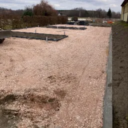 Przygotowany teren pod budowę, podsypka z kruszywa, widoczne obrzeża i słupki, w tle budynek gospodarczy i drzewa.