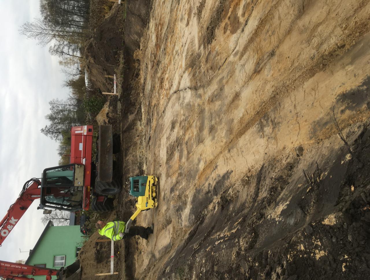 Robotnik w jaskrawej kamizelce obsługuje zagęszczarkę gruntu na nierównym, ziemistym terenie, obok czerwonego koparko-ładowarki O&K, z zielonym budynkiem i drzewami w tle.