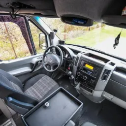 Wnętrze busa marki Mercedes z widocznym kokpitem, kierownicą i fotelami w kratę, z zawieszką zapachową na desce rozdzielczej.