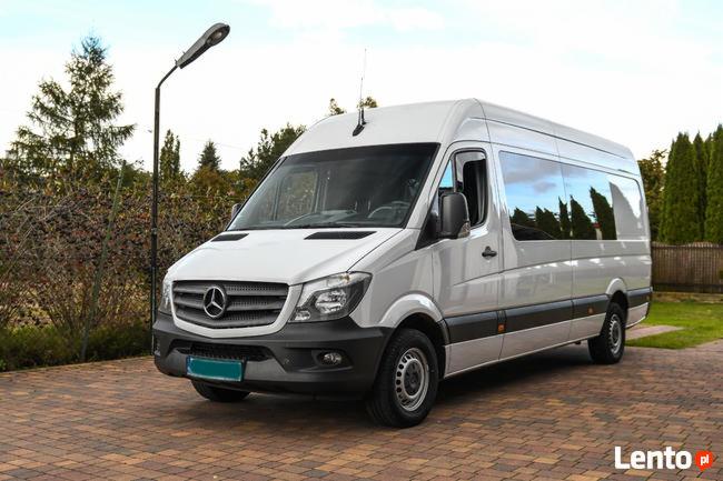 Biały bus Mercedes-Benz Sprinter z przyciemnianymi szybami, stojący na brukowanym podjeździe, z widoczną lampą uliczną i zielenią w tle.