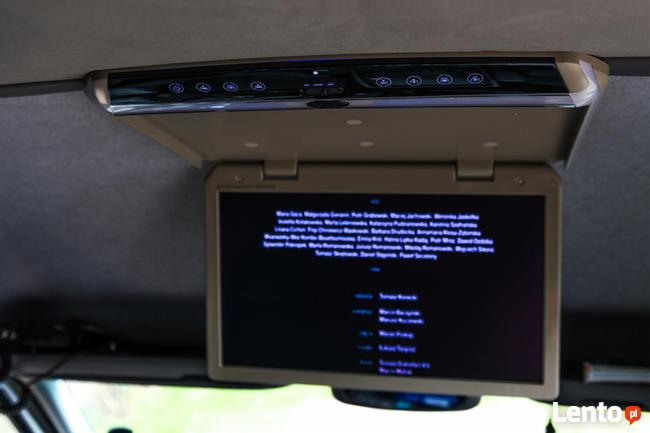 Wnętrze pojazdu z zamontowanym ekranem LCD pod sufitem, wyświetlającym listę płac.