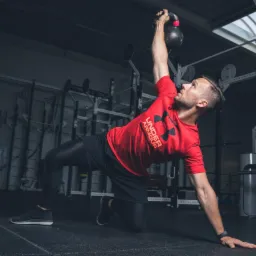Mężczyzna w czerwonej koszulce Under Armour wykonuje ćwiczenie z kettlem na siłowni, opierając się na jednej ręce i kolanie.