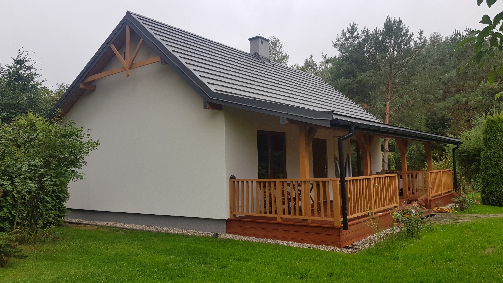 Parterowy dom z drewnianym tarasem i szarą dachówką, widok z boku na tle zieleni.