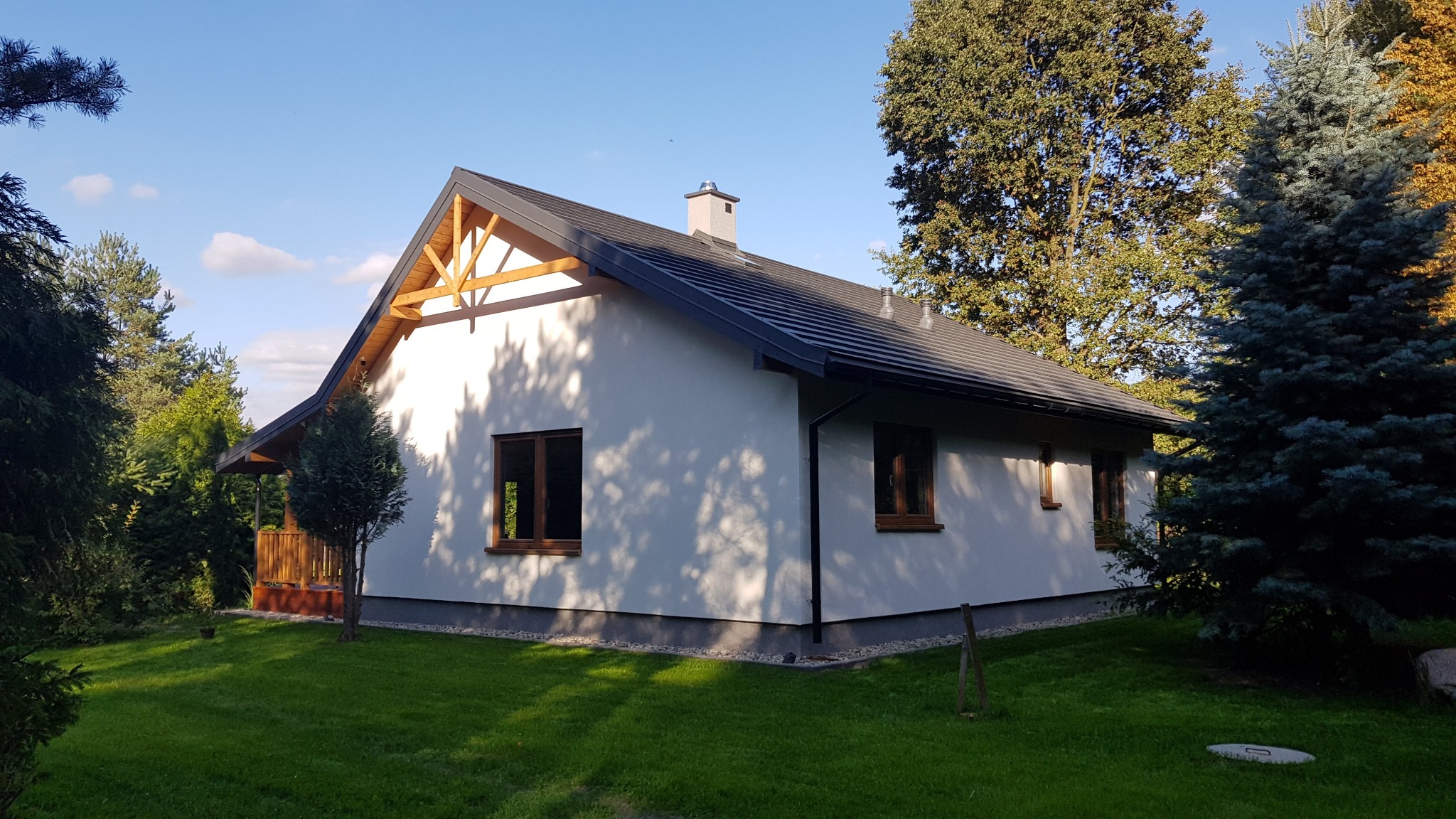 Parterowy dom jednorodzinny z białymi ścianami i ciemną dachówką, drewnianą konstrukcją pod dachem i małym tarasem, otoczony zielonym trawnikiem i drzewami w słoneczny dzień.