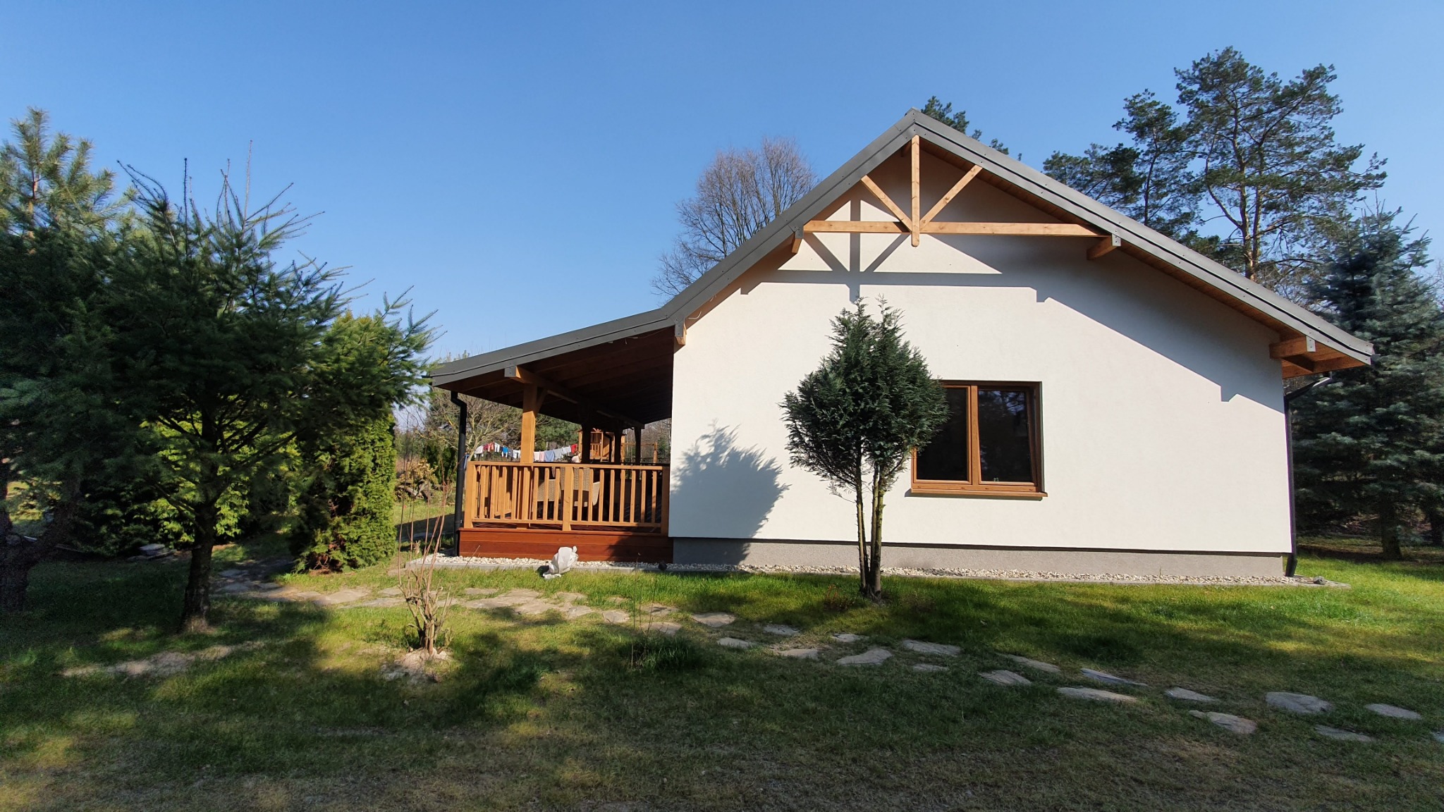 Nowoczesny dom jednorodzinny z drewnianym tarasem w Poświętnem, wykończony w jasnej kolorystyce, z widocznymi elementami konstrukcji dachu i małym drzewkiem iglastym przed budynkiem.