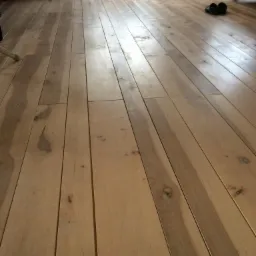 Jasne, drewniane panele podłogowe ułożone wzdłuż pomieszczenia, z widocznymi słojami i naturalnymi niedoskonałościami drewna, w tle fragment grzejnika i krzesła.