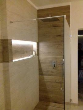 Nowoczesna kabina prysznicowa z przeszkloną ścianą, wykończona płytkami imitującymi drewno i beżowymi płytkami z wnęką oświetloną LED, widoczna bateria prysznicowa.