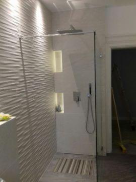 Nowoczesna kabina prysznicowa z szklaną ścianką i deszczownicą, ściana pokryta dekoracyjnymi płytkami 3D o falistej strukturze, wbudowane półki z oświetleniem punktowym, podłoga z drewnianymi...