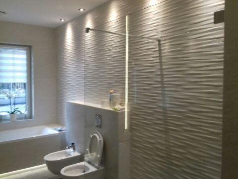 Nowoczesna łazienka z teksturowanymi płytkami ściennymi, szklaną kabiną prysznicową, wiszącą toaletą i bidetem oraz oknem z żaluzjami.