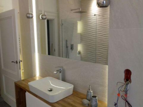 Nowoczesna łazienka z podświetlanym lustrem, umywalką nablatową na drewnianym blacie, minimalistyczną armaturą i odsłoniętymi przewodami elektrycznymi w ścianie.