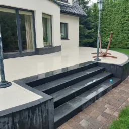 Eleganckie schody z ciemnej kostki prowadzące na jasny taras z kostki, obok domu z dużymi oknami i ogrodem. Widoczny fragment lampy i hamaka.