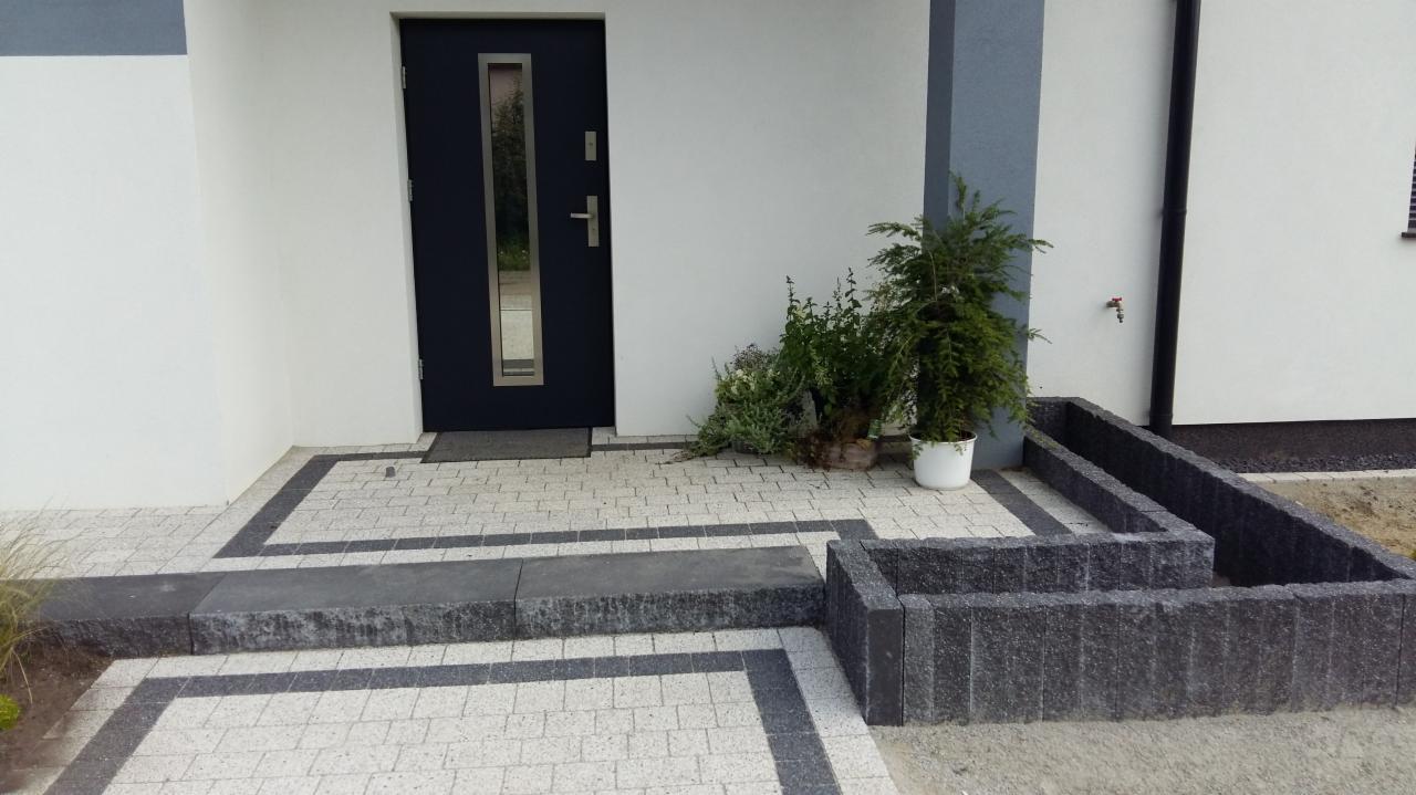 Wykończone wejście do domu z jasnej kostki brukowej z ciemnymi obramowaniami, schodkiem i rabatą z ciemnych bloczków betonowych, obok drzwi wejściowych widoczne rośliny w donicach.
