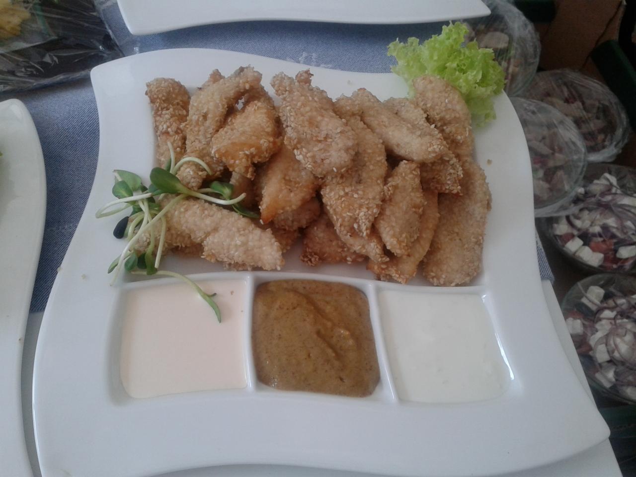 Porcja panierowanych filetów z kurczaka posypanych sezamem, podana na białym talerzu z trzema sosami (biały, musztardowy i biały) oraz dekoracją z zielonej sałaty i kiełków.