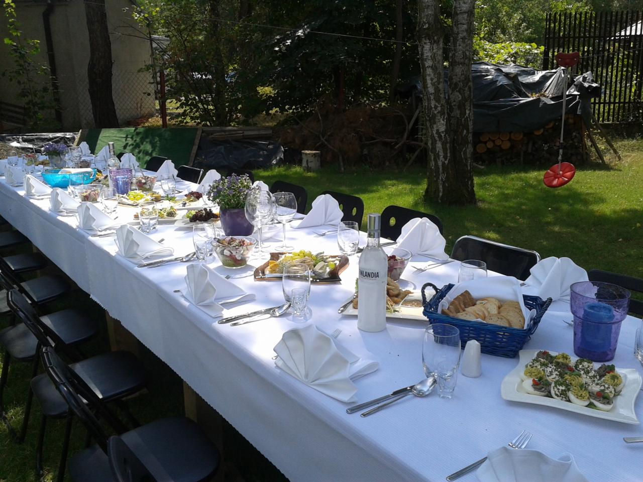 Długi stół cateringowy na zewnątrz, nakryty białym obrusem, z potrawami, napojami i dekoracjami, ustawiony na trawniku z drzewami w tle, z widocznym fragmentem huśtawki.