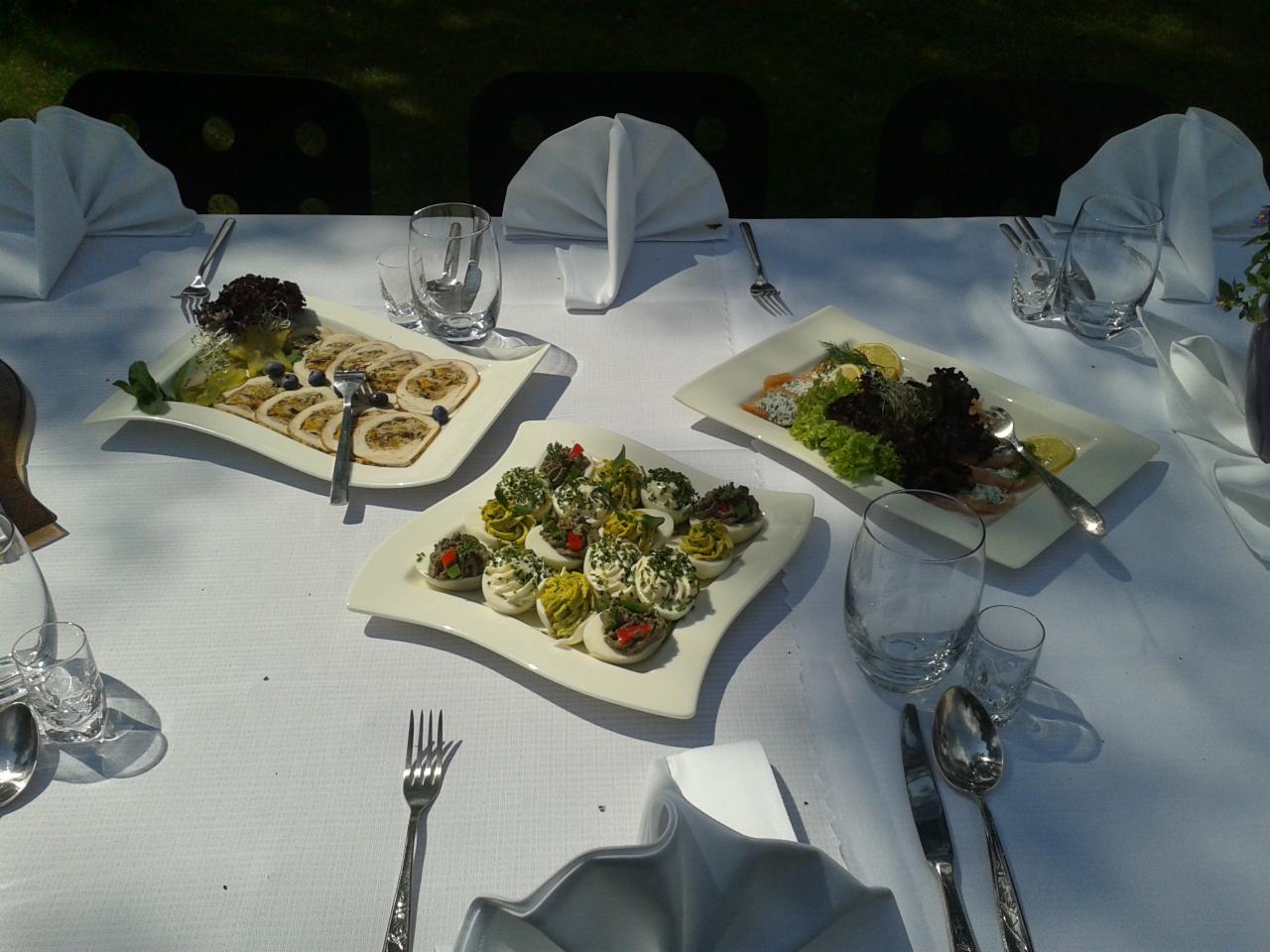 Elegancko zastawiony stół na zewnątrz z trzema różnymi rodzajami przystawek na białych, asymetrycznych talerzach: roladki mięsne z borówkami, jajka faszerowane z ziołami i papryką oraz wędzony...