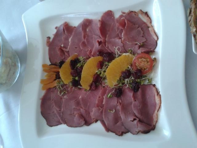 Dekoracyjnie ułożone plastry wędliny na kwadratowym białym talerzu, ozdobione pomarańczami, żurawiną, kiełkami i plasterkiem pomidora.