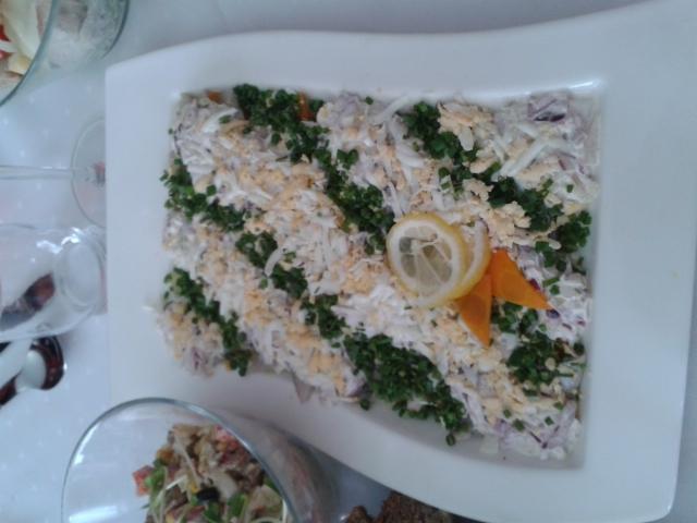 Śledź pod pierzynką na białym prostokątnym talerzu, udekorowany plasterkiem cytryny i marchewką, posypany koperkiem i startym jajkiem, widoczne inne sałatki w szklanych misach w tle