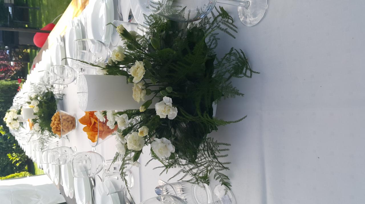 Długi stół z białym obrusem, nakryty białymi talerzami i kieliszkami, udekorowany kompozycjami kwiatowymi z białych goździków i zieleni, z centralnie umieszczoną grubą białą świecą oraz przekąskami...