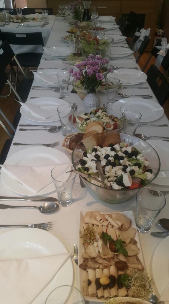 Ustawiony stół z białym obrusem, nakryciami, szklankami i różnorodnymi potrawami, w tym sałatkami, wędlinami i kwiatami w wazonach.